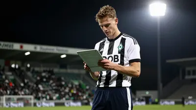 A young Werder Bremen coach in a white and black striped shirt and navy tracksuit pants studies a detailed scouting...