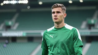 A young, athletic Werderaner U21 player, mid-20s, in a plain green and white training top, stands pitchside at the...